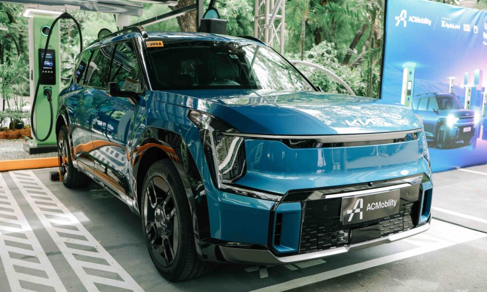 A blue Kia EV9 electric vehicle is parked at an ACMobility fast charging station in Makati. The vehicle is plugged into a green EV charger, with ACMobility branding and promotional banners visible in the background. The setting is surrounded by trees and greenery, emphasizing a clean and sustainable urban environment.