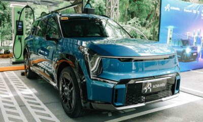 A blue Kia EV9 electric vehicle is parked at an ACMobility fast charging station in Makati. The vehicle is plugged into a green EV charger, with ACMobility branding and promotional banners visible in the background. The setting is surrounded by trees and greenery, emphasizing a clean and sustainable urban environment.