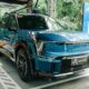 A blue Kia EV9 electric vehicle is parked at an ACMobility fast charging station in Makati. The vehicle is plugged into a green EV charger, with ACMobility branding and promotional banners visible in the background. The setting is surrounded by trees and greenery, emphasizing a clean and sustainable urban environment.