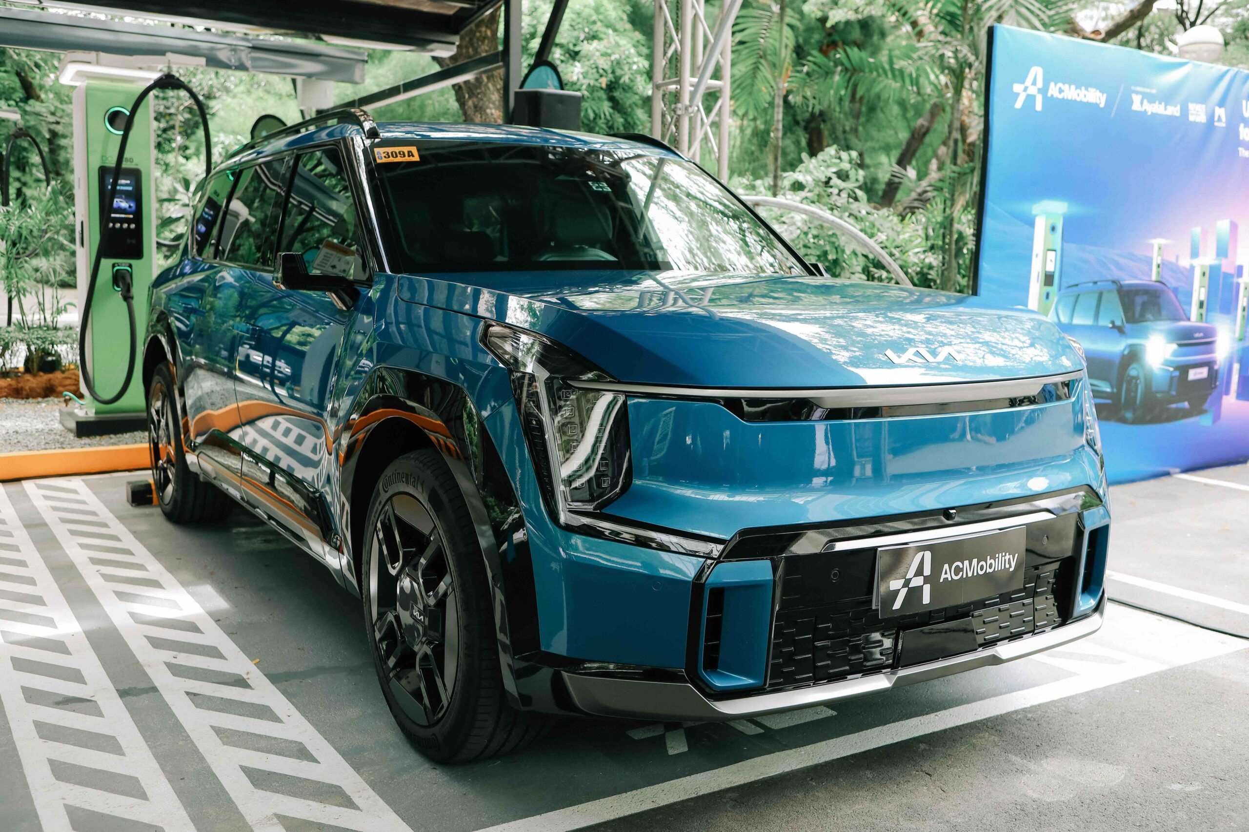 A blue Kia EV9 electric vehicle is parked at an ACMobility fast charging station in Makati. The vehicle is plugged into a green EV charger, with ACMobility branding and promotional banners visible in the background. The setting is surrounded by trees and greenery, emphasizing a clean and sustainable urban environment.