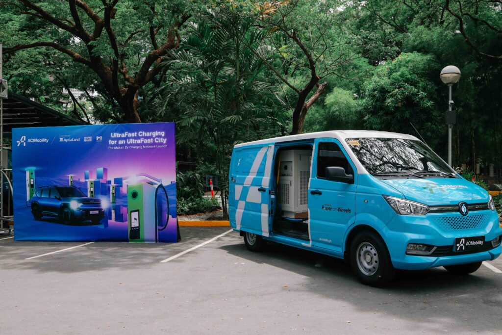 A light blue ACMobility Power-on-Wheels van is parked with its side door open, revealing EV charging equipment inside. Beside it stands a promotional display highlighting ACMobility’s Makati EV Charging Network launch, featuring slogans like “UltraFast Charging for an UltraFast City.” The scene is set in a tree-lined urban area, underscoring the eco-friendly theme of mobile fast-charging infrastructure.