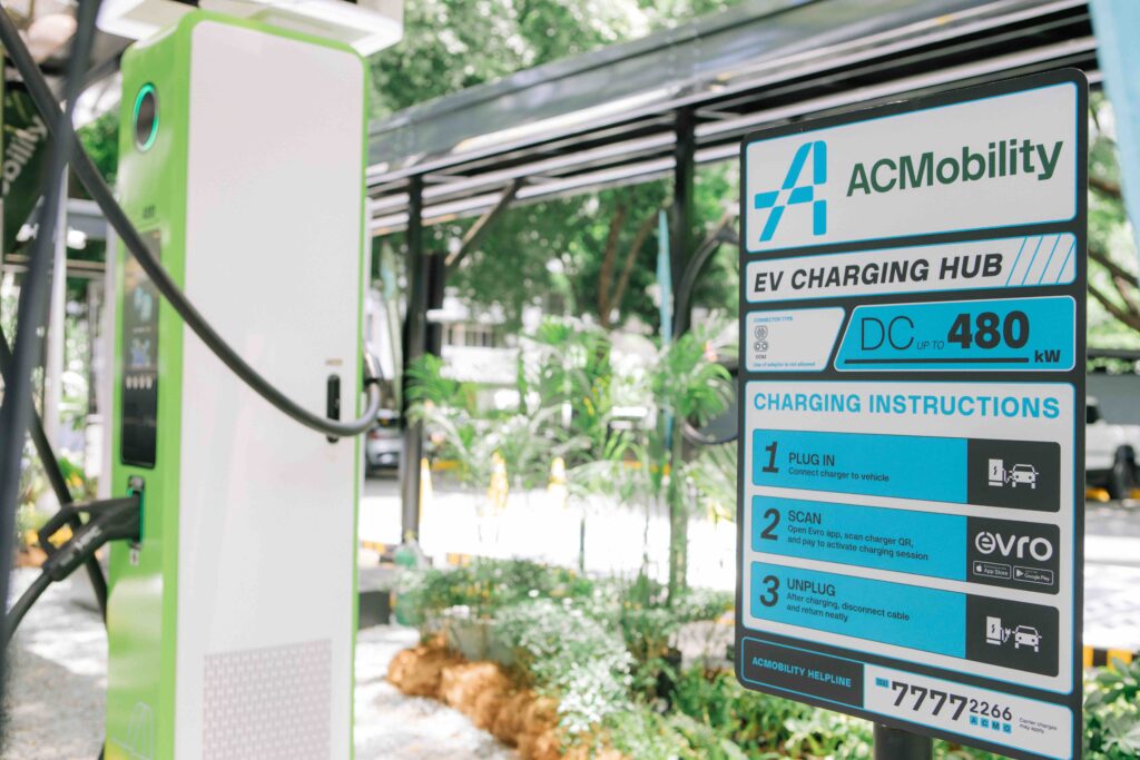 Close-up of a green and white 480 kW ACMobility DC fast EV charger beside a clear instruction board labeled “EV Charging Hub.” The signage outlines the steps for plugging in, scanning the Evro app, and unplugging after charging. The setting is outdoors, with greenery and a canopy-covered charging area in the background, showcasing a modern, user-friendly charging setup.