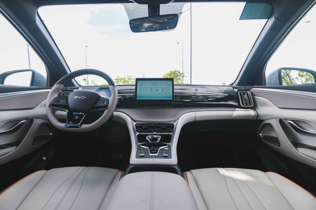 Interior view of the BYD Sealion 5 DM-i showing a minimalist dashboard with a rotating touchscreen, sleek steering wheel with controls, and two-tone leather seats.

