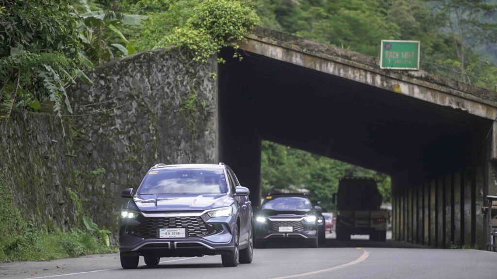 Two BYD Sealion 5 DM-i units drive out of a mountain rock shed tunnel, surrounded by thick greenery on a winding road in Luzon, Philippines.