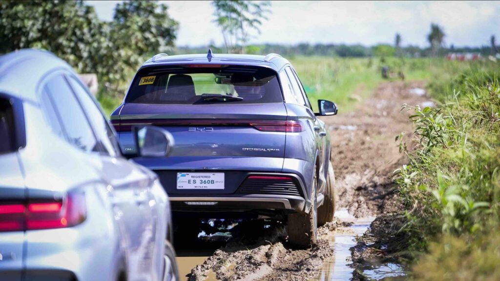 A rear view of the BYD Sealion 5 DM-i navigating through a muddy, narrow rural path with another electrified vehicle closely following behind.
