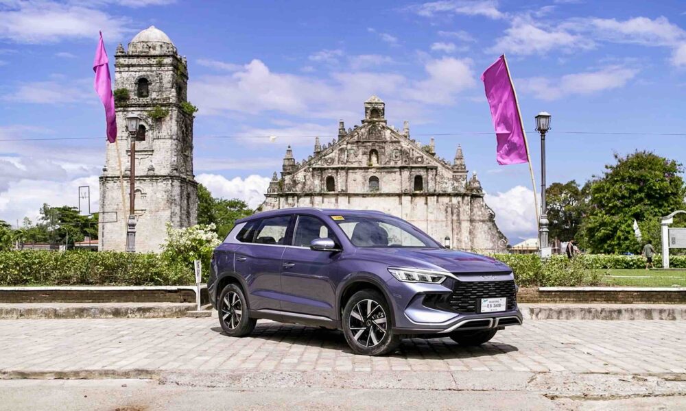 A BYD Sealion 5 DM-i in Deep Sea Blue is parked in front of the historic Paoay Church in Ilocos Norte, with pink flags waving in the background under a bright blue sky.