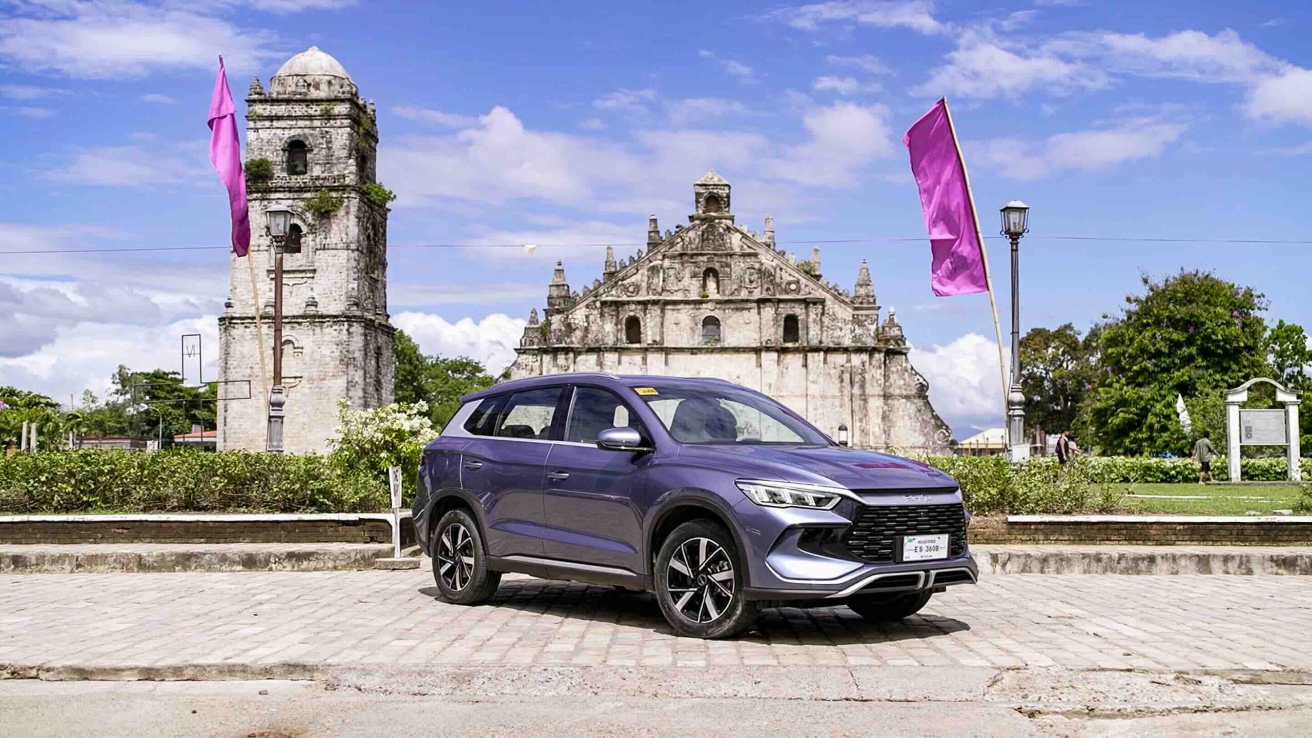 A BYD Sealion 5 DM-i in Deep Sea Blue is parked in front of the historic Paoay Church in Ilocos Norte, with pink flags waving in the background under a bright blue sky.