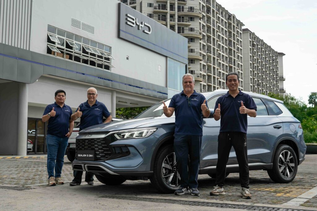 Four men in matching navy blue shirts give thumbs-up poses in front of the BYD Sealion 5 DM-i, parked outside a BYD dealership with a high-rise condo building in the background.