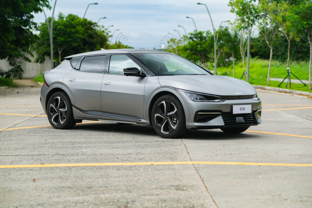 A sleek matte gray Kia EV6 is parked in an open lot lined with yellow markings and surrounded by trees and lamp posts, highlighting its aerodynamic profile and modern wheels.