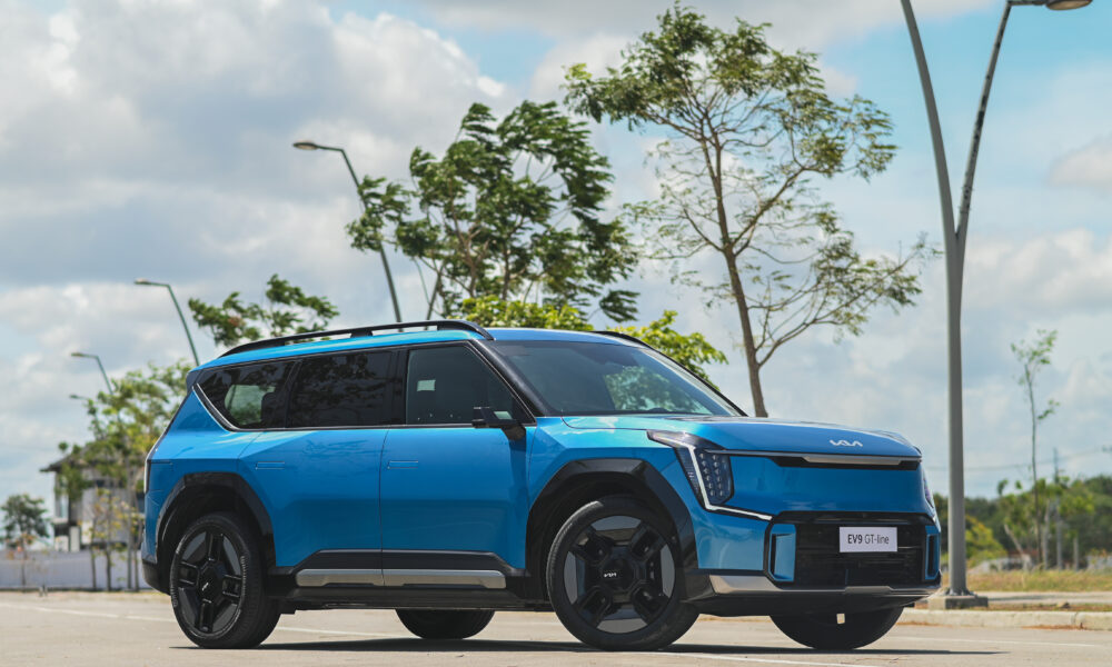 A bright blue Kia EV9 GT-Line is parked on a sunny street, surrounded by trees and street lamps, showcasing its bold design and black alloy wheels.