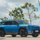 A bright blue Kia EV9 GT-Line is parked on a sunny street, surrounded by trees and street lamps, showcasing its bold design and black alloy wheels.