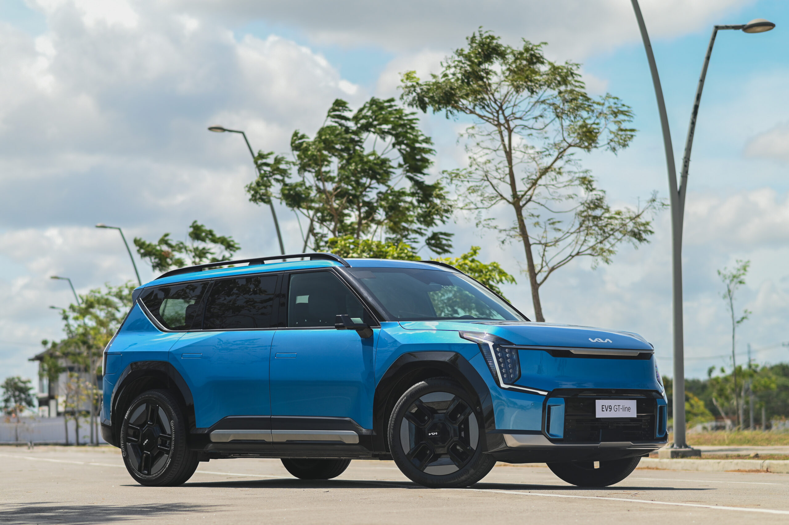 A bright blue Kia EV9 GT-Line is parked on a sunny street, surrounded by trees and street lamps, showcasing its bold design and black alloy wheels.
