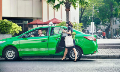 A bright green electric taxi is parked by the sidewalk in a city area, with a smiling male driver inside. A woman carrying a white tote bag and fresh produce is about to get into the back seat. The scene is framed by palm trees, red umbrellas, and urban buildings, suggesting a clean and modern commute.