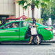 A bright green electric taxi is parked by the sidewalk in a city area, with a smiling male driver inside. A woman carrying a white tote bag and fresh produce is about to get into the back seat. The scene is framed by palm trees, red umbrellas, and urban buildings, suggesting a clean and modern commute.