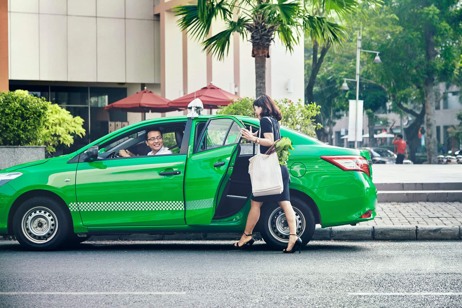 A bright green electric taxi is parked by the sidewalk in a city area, with a smiling male driver inside. A woman carrying a white tote bag and fresh produce is about to get into the back seat. The scene is framed by palm trees, red umbrellas, and urban buildings, suggesting a clean and modern commute.