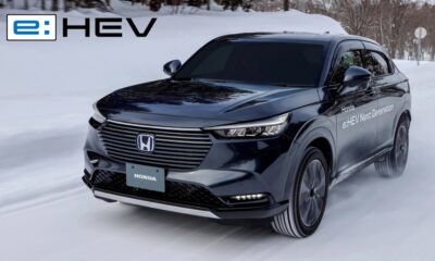 A sleek black Honda e:HEV Next Generation hybrid SUV driving confidently on a snowy road, showcasing its electric all-wheel drive capability. The vehicle kicks up snow as it maintains traction, with the e:HEV logo in the upper left corner of the image.