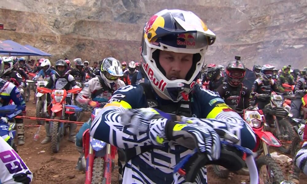 Manuel Lettenbichler, wearing a Red Bull helmet and racing gear, prepares at the starting line of the 2025 Red Bull Erzbergrodeo with a focused expression. Behind him, a sea of fellow riders on dirt bikes waits in anticipation amid the rugged quarry backdrop of Eisenerz, Austria.