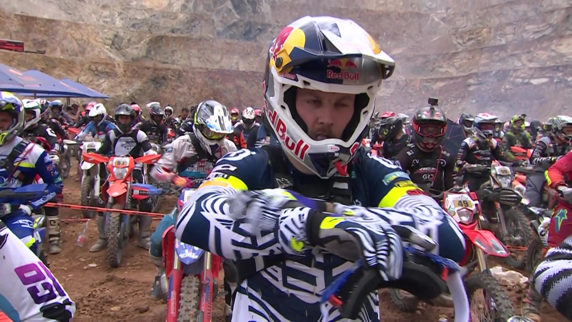 Manuel Lettenbichler, wearing a Red Bull helmet and racing gear, prepares at the starting line of the 2025 Red Bull Erzbergrodeo with a focused expression. Behind him, a sea of fellow riders on dirt bikes waits in anticipation amid the rugged quarry backdrop of Eisenerz, Austria.