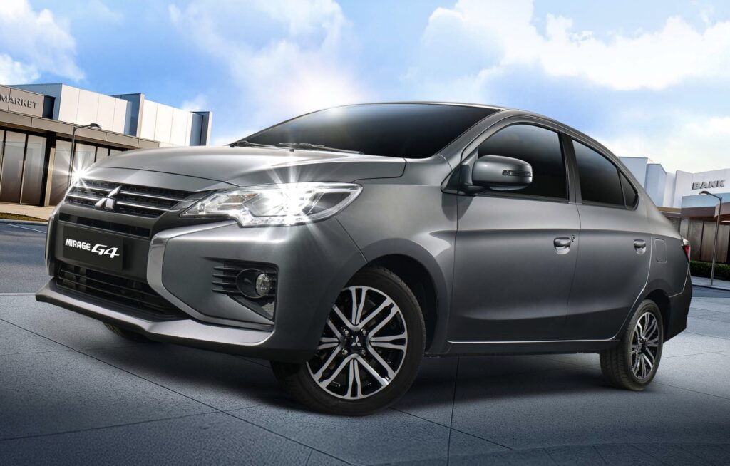 A sleek gray Mitsubishi Mirage G4 sedan is parked on a paved surface in front of modern buildings with signs reading "MARKET" and "BANK." The car features bright headlights and stylish alloy wheels, set against a backdrop of a clear blue sky with scattered clouds.