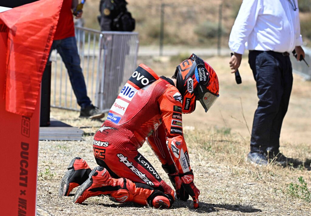 Marc Marquez kneels on the gravel beside the track in full red Ducati racing gear and helmet, appearing emotional and reflective after the Aragon MotoGP, with team staff and officials in the background.
