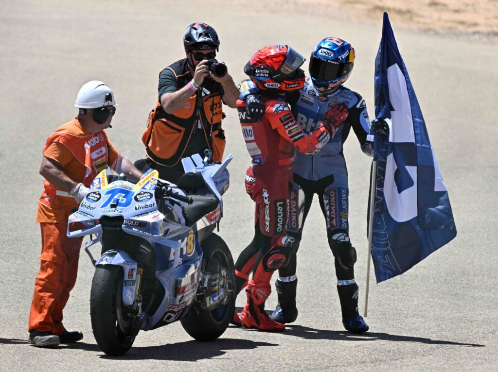 Marc and Alex Marquez share an emotional helmeted embrace on the track after their 1-2 finish at the Aragon MotoGP, with Alex holding a large flag and race officials standing nearby next to his number 73 bike. A photographer captures the celebratory moment.