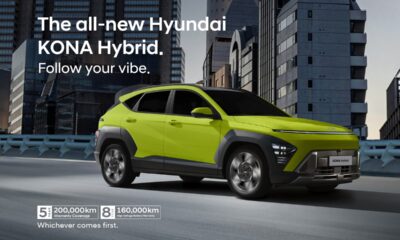 A vibrant lime-green Hyundai KONA Hybrid drives along an elevated city road surrounded by tall, modern buildings at dusk. The car’s futuristic design is highlighted by LED headlights, bold lines, and contrasting black wheel arches, capturing its dynamic urban character.