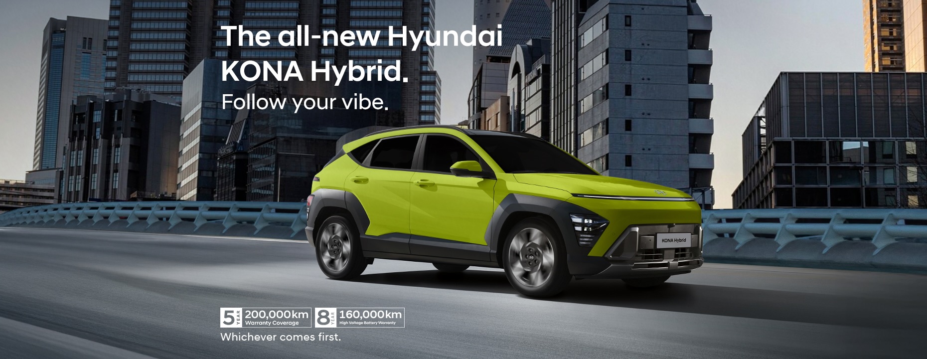 A vibrant lime-green Hyundai KONA Hybrid drives along an elevated city road surrounded by tall, modern buildings at dusk. The car’s futuristic design is highlighted by LED headlights, bold lines, and contrasting black wheel arches, capturing its dynamic urban character.