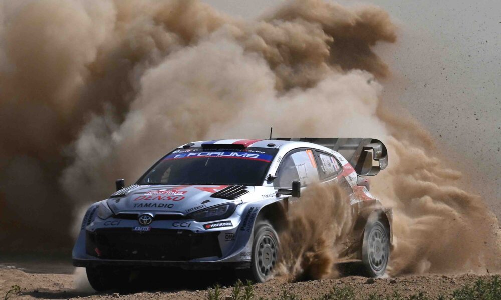 A Toyota GR Yaris Rally1 kicks up a massive cloud of dust as it powers through a gravel corner during the 2024 Rally of Sardinia. The car, bearing Sebastien Ogier’s number and Red Bull branding, displays dirt and grit from the intense competition.