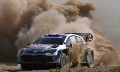A Toyota GR Yaris Rally1 kicks up a massive cloud of dust as it powers through a gravel corner during the 2024 Rally of Sardinia. The car, bearing Sebastien Ogier’s number and Red Bull branding, displays dirt and grit from the intense competition.