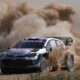 A Toyota GR Yaris Rally1 kicks up a massive cloud of dust as it powers through a gravel corner during the 2024 Rally of Sardinia. The car, bearing Sebastien Ogier’s number and Red Bull branding, displays dirt and grit from the intense competition.