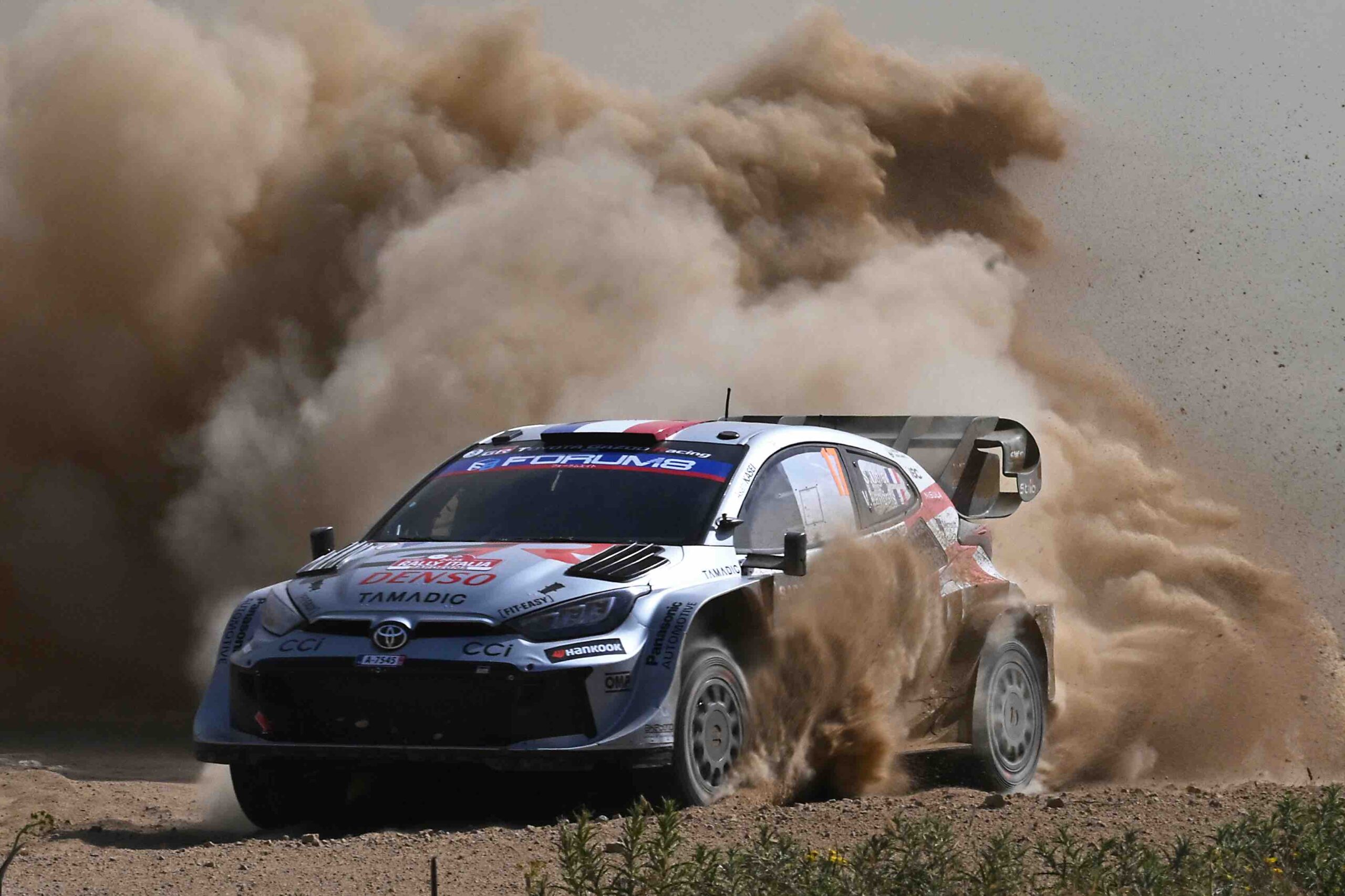 A Toyota GR Yaris Rally1 kicks up a massive cloud of dust as it powers through a gravel corner during the 2024 Rally of Sardinia. The car, bearing Sebastien Ogier’s number and Red Bull branding, displays dirt and grit from the intense competition.