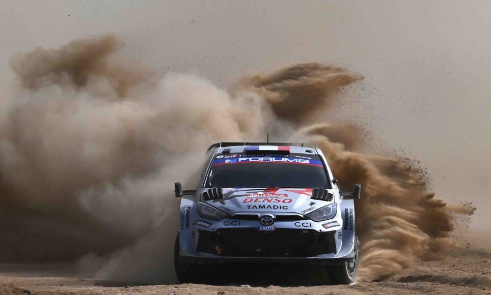 A Toyota GR Yaris Rally1 kicks up a massive cloud of dust as it powers through a gravel corner during the 2024 Rally of Sardinia. The car, bearing Sebastien Ogier’s number and Red Bull branding, displays dirt and grit from the intense competition.