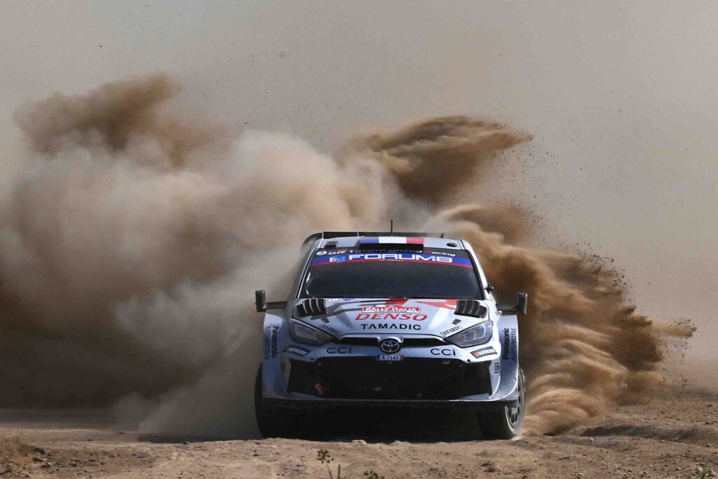A Toyota GR Yaris Rally1 kicks up a massive cloud of dust as it powers through a gravel corner during the 2024 Rally of Sardinia. The car, bearing Sebastien Ogier’s number and Red Bull branding, displays dirt and grit from the intense competition.