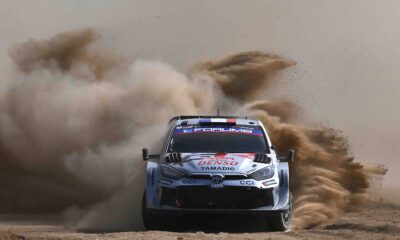 A Toyota GR Yaris Rally1 kicks up a massive cloud of dust as it powers through a gravel corner during the 2024 Rally of Sardinia. The car, bearing Sebastien Ogier’s number and Red Bull branding, displays dirt and grit from the intense competition.