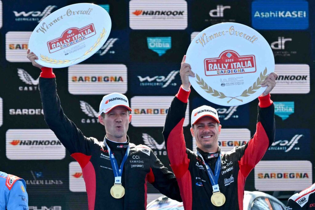 Sebastien Ogier and co-driver Vincent Landais celebrate on the podium after winning the 2024 Rally Italia Sardegna. Both are wearing black and red Toyota Gazoo Racing suits and Hankook caps, proudly holding up circular "Winner Overall" rally trophies and wearing gold medals.