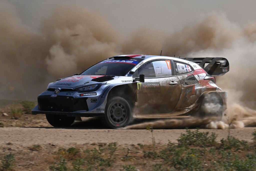 A Toyota GR Yaris Rally1 kicks up a massive cloud of dust as it powers through a gravel corner during the 2024 Rally of Sardinia. The car, bearing Sebastien Ogier’s number and Red Bull branding, displays dirt and grit from the intense competition.