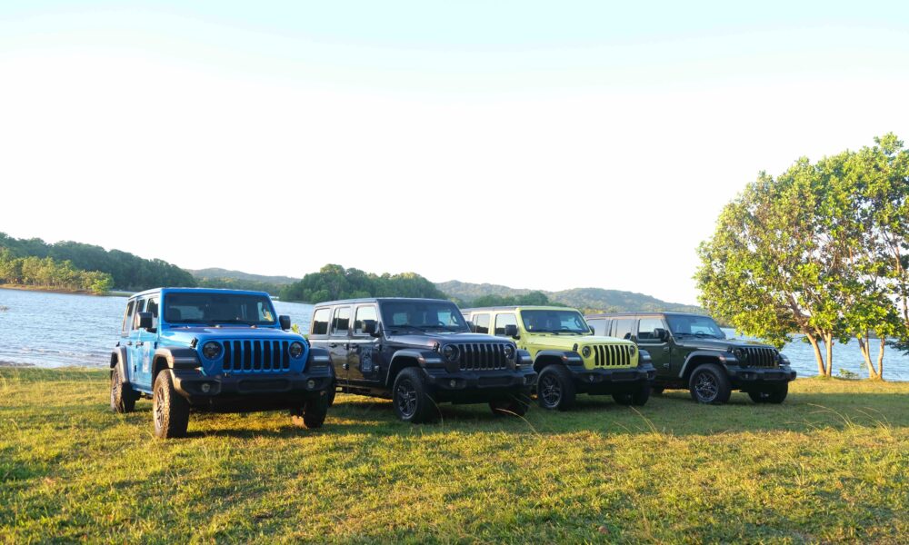 A lineup of colorful Jeep® Wrangler 4-Door models parked on a grassy field beside a lake, with hills and trees in the background under a clear sky.