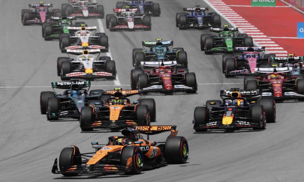 A pack of Formula One cars battles for position at the start of a Grand Prix race. Leading the group are McLaren, Red Bull, and Mercedes cars, followed closely by Ferrari, Aston Martin, and other teams. The cars are on a wide tarmac track with red and white curbs, under bright daylight.