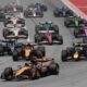 A pack of Formula One cars battles for position at the start of a Grand Prix race. Leading the group are McLaren, Red Bull, and Mercedes cars, followed closely by Ferrari, Aston Martin, and other teams. The cars are on a wide tarmac track with red and white curbs, under bright daylight.