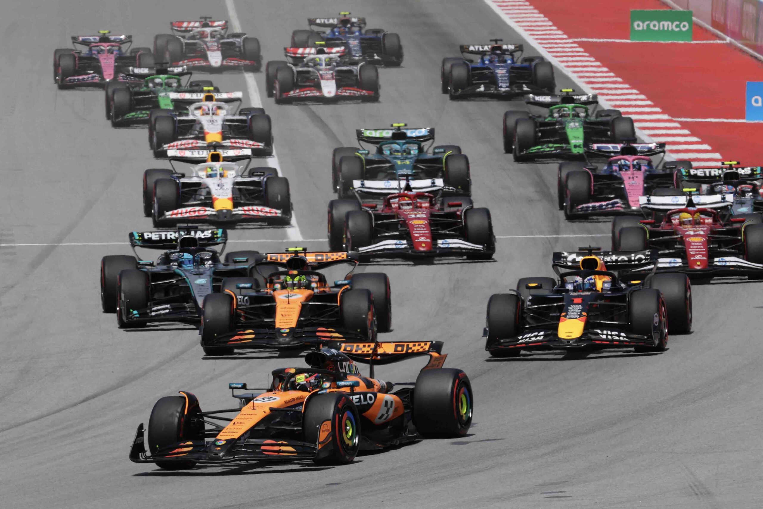 A pack of Formula One cars battles for position at the start of a Grand Prix race. Leading the group are McLaren, Red Bull, and Mercedes cars, followed closely by Ferrari, Aston Martin, and other teams. The cars are on a wide tarmac track with red and white curbs, under bright daylight.