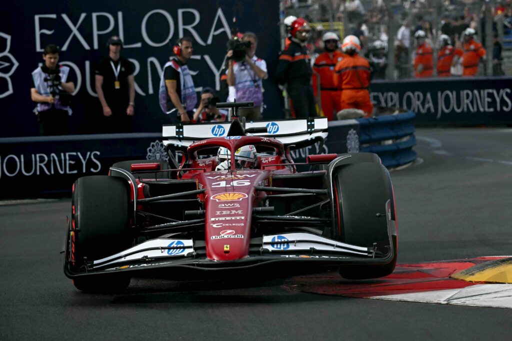A Ferrari Formula One car, driven by Charles Leclerc (#16), speeds through a tight corner on a street circuit. The front wheels are lifted slightly off the ground as the car clips the red and yellow curb. Track marshals and photographers watch from behind safety barriers.

