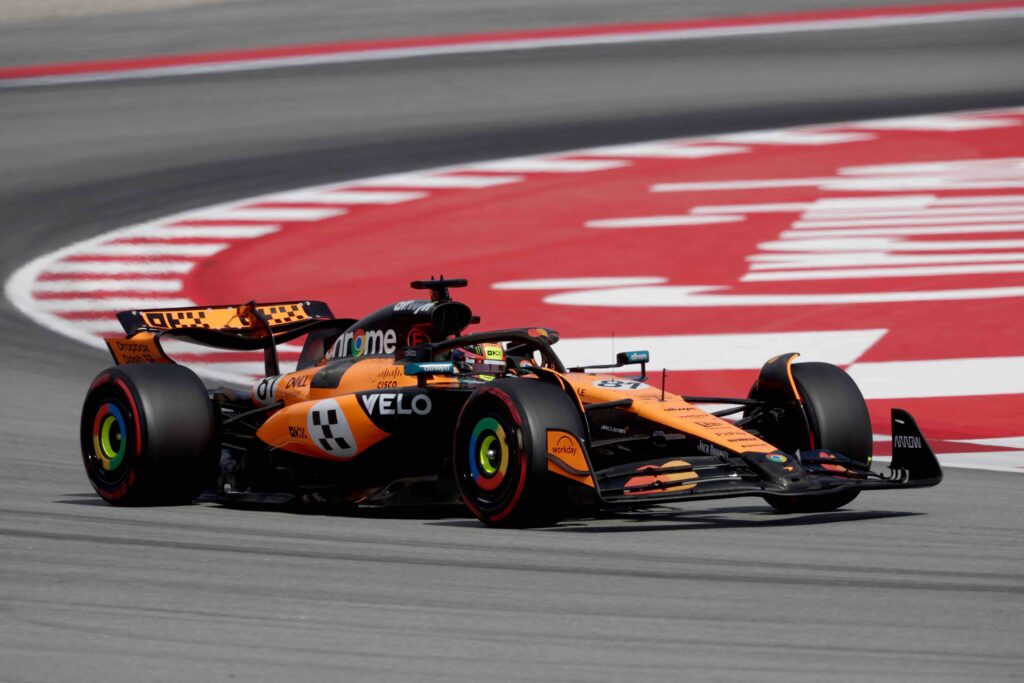A McLaren Formula One car, painted in striking orange and black, navigates a high-speed corner on a racetrack with red and white curbs. The car is low to the ground, hugging the apex with precision.

