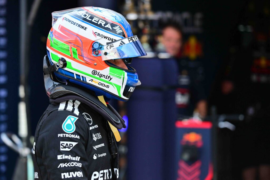 A Formula One driver wearing a colorful helmet and a black Mercedes-AMG Petronas racing suit walks through the pit lane. The helmet features vibrant red, green, and blue accents along with sponsor logos. The background shows blurred team personnel and garage equipment.
