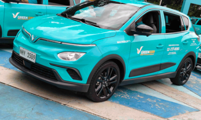 A brightly colored Green GSM electric taxi, a VinFast Nerio EV, is parked at a launch event in Quezon City. The car features a sleek cyan body with black rims and the Green GSM hotline visibly printed on the door. A driver sits inside while another e-taxi is seen beside it.