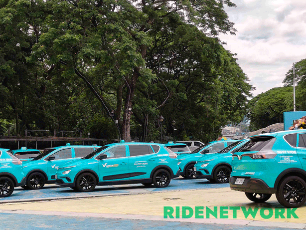 A fleet of Green GSM electric taxis lines up at Quezon Memorial Circle, with cyan-colored VinFast Nerio EVs ready for launch under lush green trees. The RIDENETWORK watermark is placed at the lower right corner of the image.
