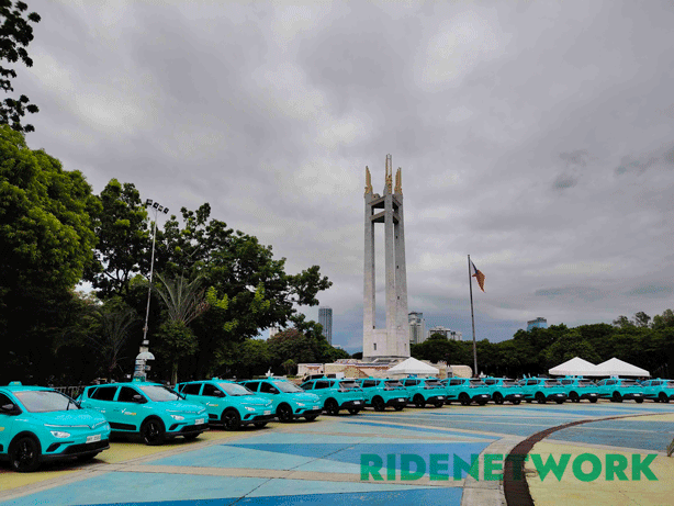 A striking lineup of Green GSM electric taxis encircles the Quezon Memorial Shrine at Quezon Memorial Circle. The fleet of bright cyan VinFast Nerio EVs stands in contrast to the cloudy sky and national monument. The RIDENETWORK watermark is visible on the pavement.