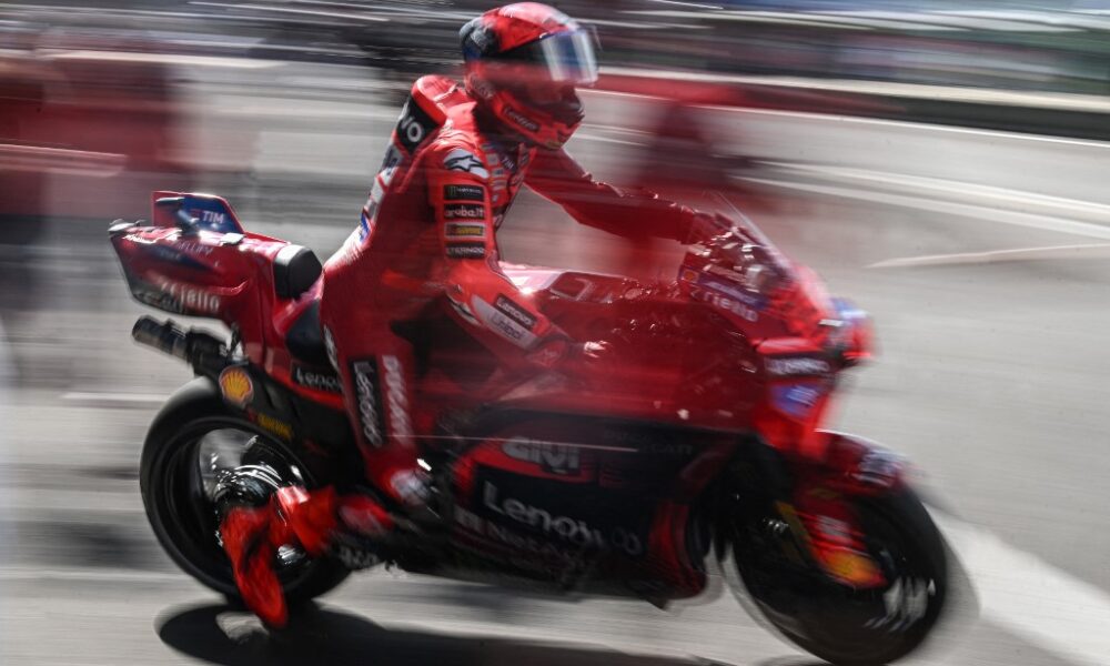 A red-clad MotoGP rider on a Ducati bike streaks past the pit lane, captured with dramatic motion blur.
