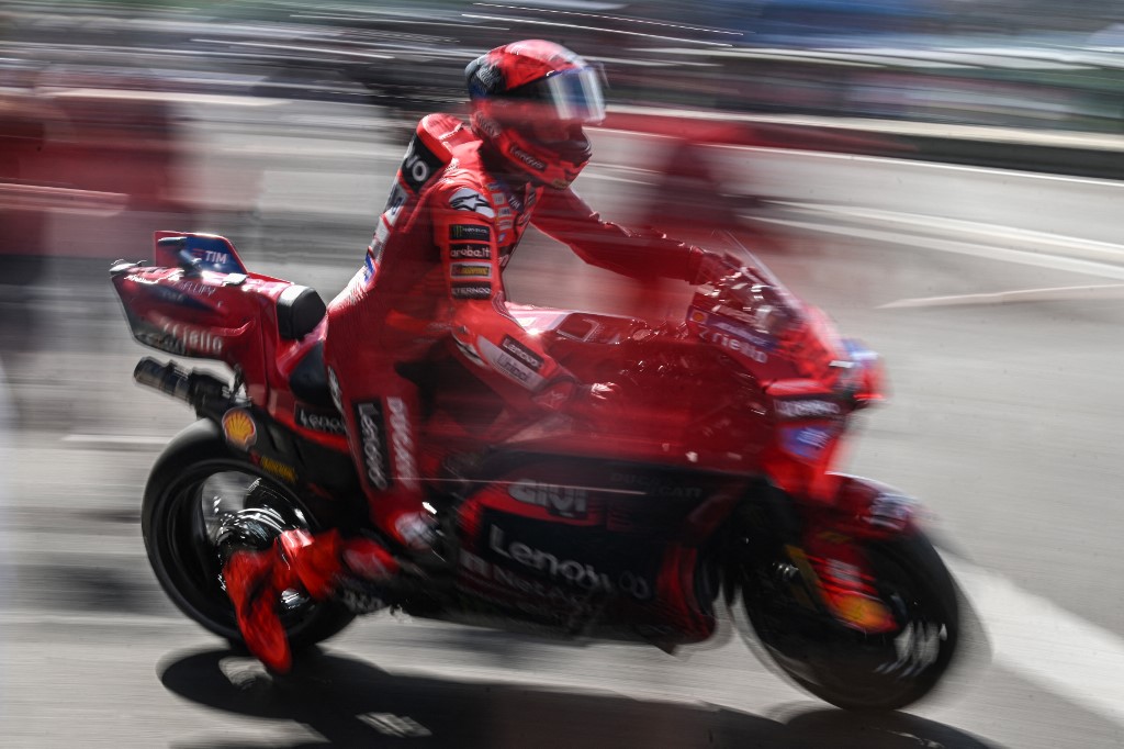 A red-clad MotoGP rider on a Ducati bike streaks past the pit lane, captured with dramatic motion blur.