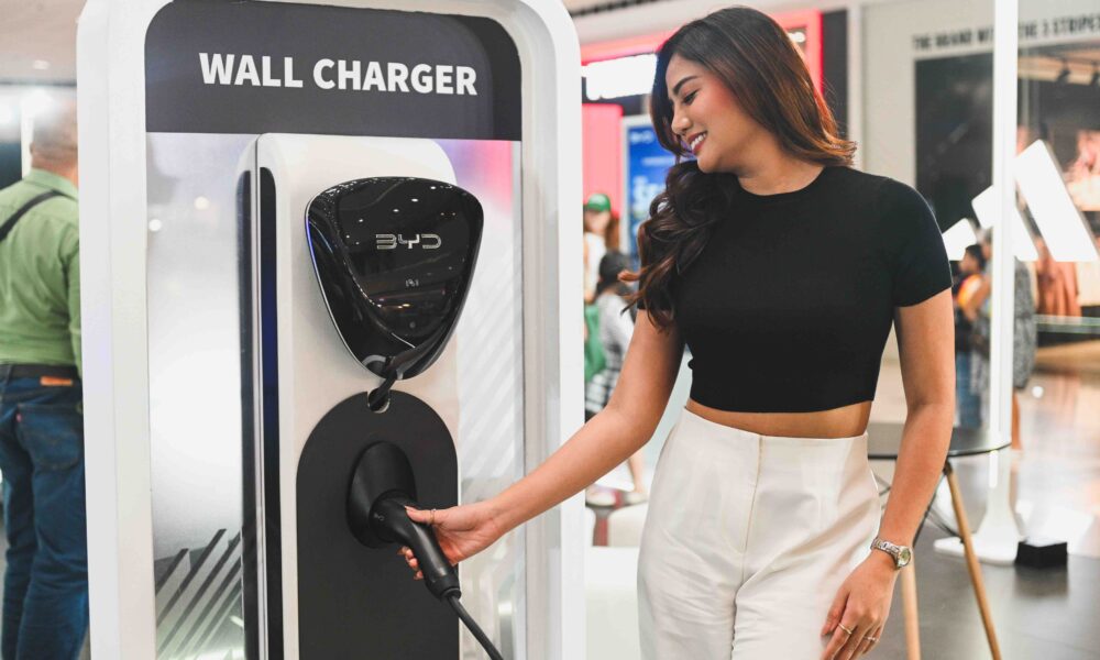 Woman smiling while holding the plug of a BYD electric vehicle wall charger at an indoor exhibit. Let me know if you need more images captioned or want to feature this on your site or socials!