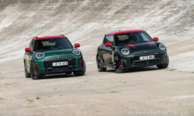 Two all-new MINI John Cooper Works models—an Aceman in Rebel Green and a 3-door Electric in Midnight Black—driving side by side on a steeply banked concrete track.